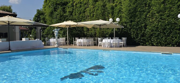 Vista diurna di una piscina esterna, con numerosi tavoli da ricevimento elegantemente apparecchiati con tovaglie bianche pulite disposti a bordo vasca. Ombrelloni da esterno offrono ombra e riparo dal sole. Sullo sfondo si estende una fitta siepe verde e curata, che delimita il giardino.