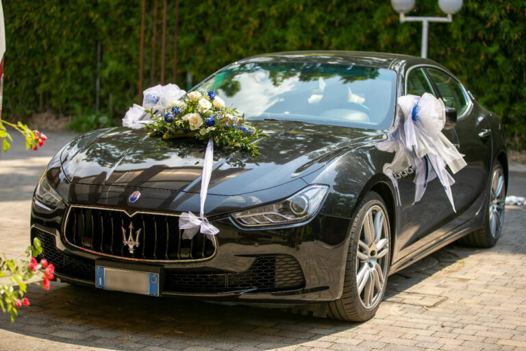 Elegante automobile di lusso berlina nera addobbata per un matrimonio, decorata con fiocchi bianchi e blu fissati agli specchietti. Un raffinato bouquet di fiori freschi è appoggiato centralmente sul cofano.