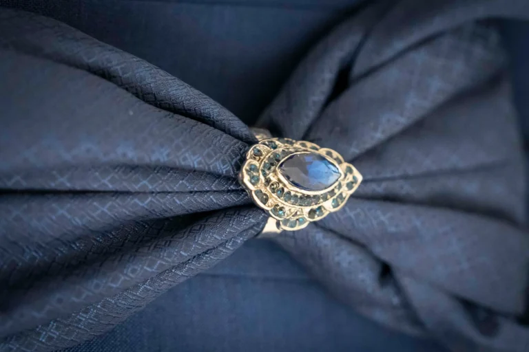 Primo piano ravvicinato che mostra un gioiello a forma di goccia con pietra preiosa blu con un intricato motivo a rilievo, adagiato e parzialmente avvolto da un tessuto in raso o seta di colore blu notte intenso