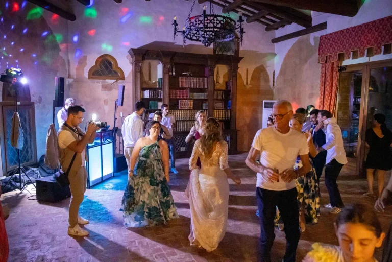 Scena dinamica di invitati a un matrimonio che ballano e festeggiano all'interno della lussuosa sala di ricevimento di una villa storica. Lo spazio è elegantemente decorato con lampadari d'epoca in cristallo e una grande libreria piena di volumi antichi. L'illuminazione ambientale, con luci soffuse e decorative (uplighting), crea un'atmosfera calda e festosa