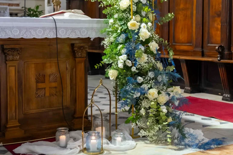 Dettaglio di un allestimento nuziale in chiesa, che mostra candele votive accese in vasi di vetro trasparente e maestosi archi floreali composti da rose e fogliame fresco, posizionati ai lati di un altare.