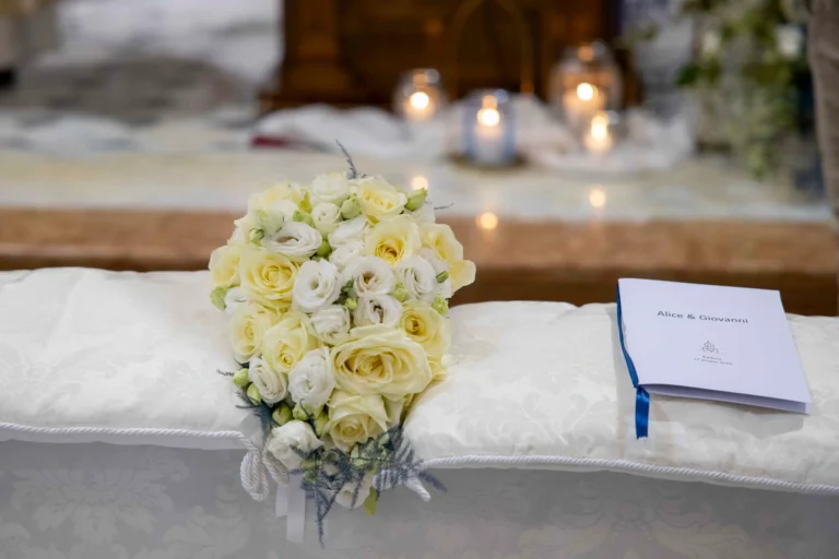 Primo piano ravvicinato di un delicato bouquet di rose fresche bianche e giallo pastello, adagiato su un morbido cuscino di un banco di chiesa. Accanto al bouquet, un elegante cartoncino decorato con nastro blu con i nomi degli sposi stampati in bella calligrafia.