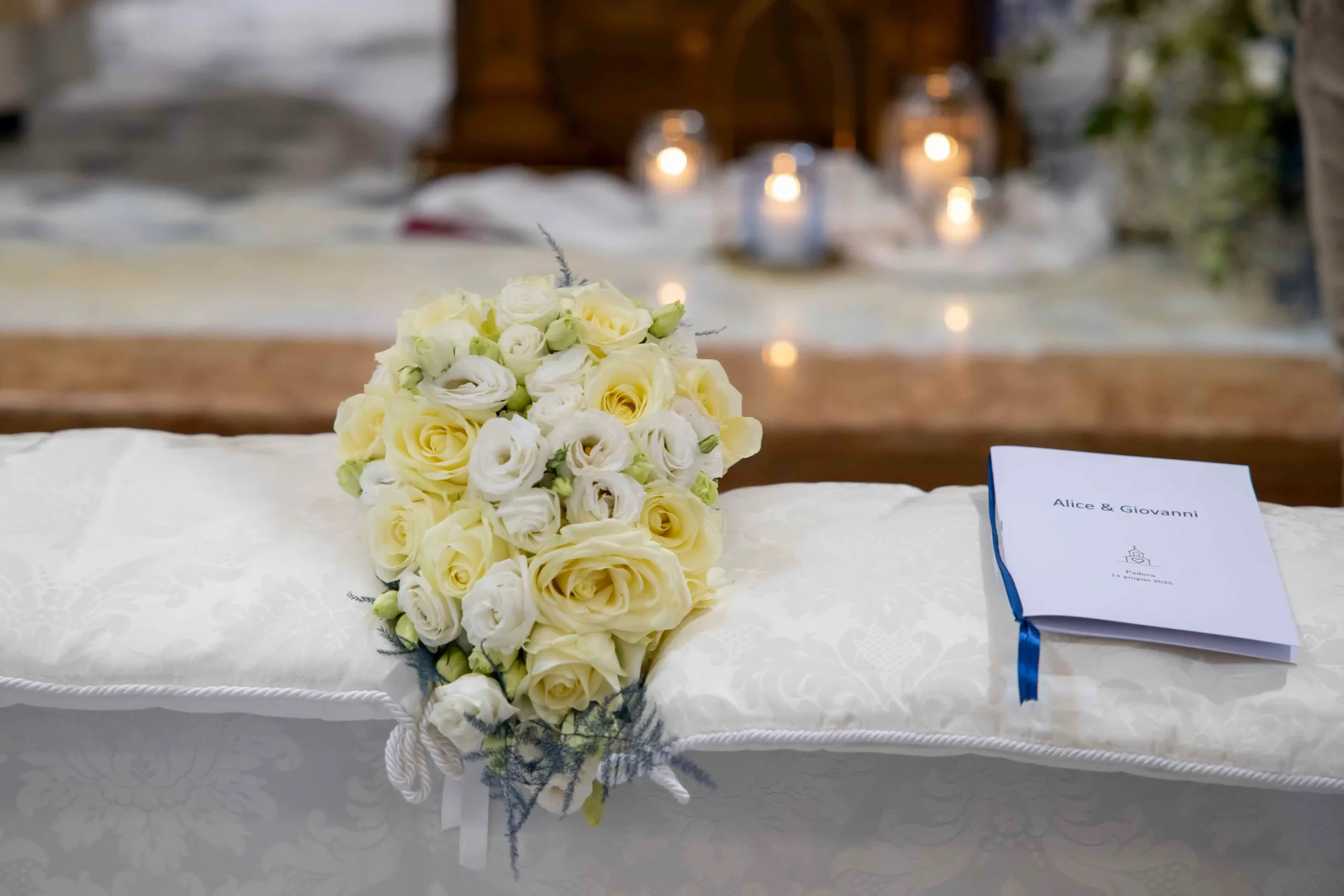 Primo piano ravvicinato di un delicato bouquet di rose fresche bianche e giallo pastello, adagiato su un morbido cuscino di un banco di chiesa. Accanto al bouquet, un elegante cartoncino decorato con nastro blu con i nomi degli sposi stampati in bella calligrafia.