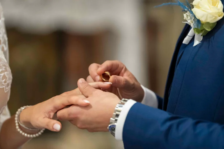 Due mani di uno sposo che mette al dito della sposa l'anello nuziale. Si vedono dettagli come accessori ai polsi e dettagli dei vestiti