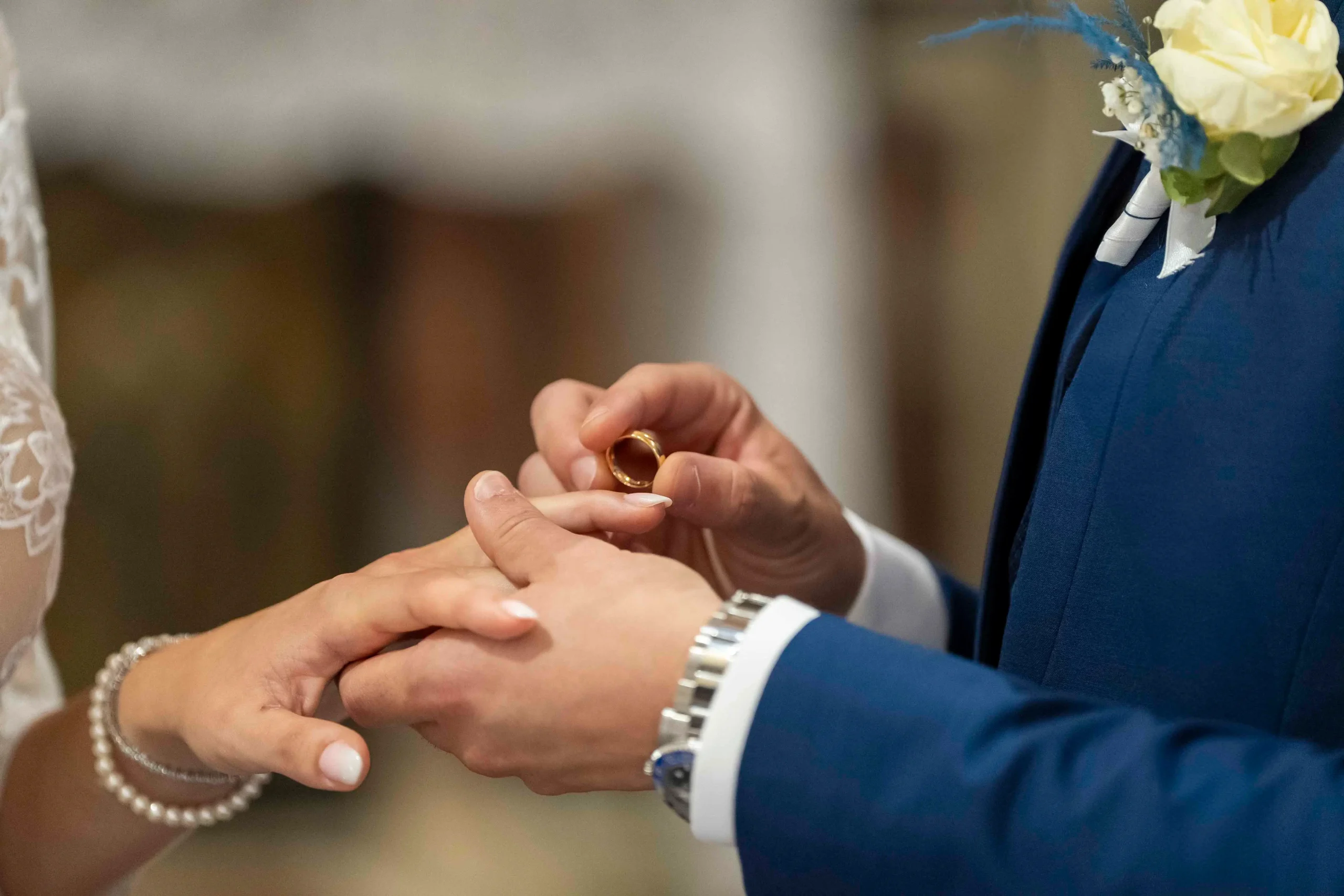 Due mani di uno sposo che mette al dito della sposa l'anello nuziale. Si vedono dettagli come accessori ai polsi e dettagli dei vestiti