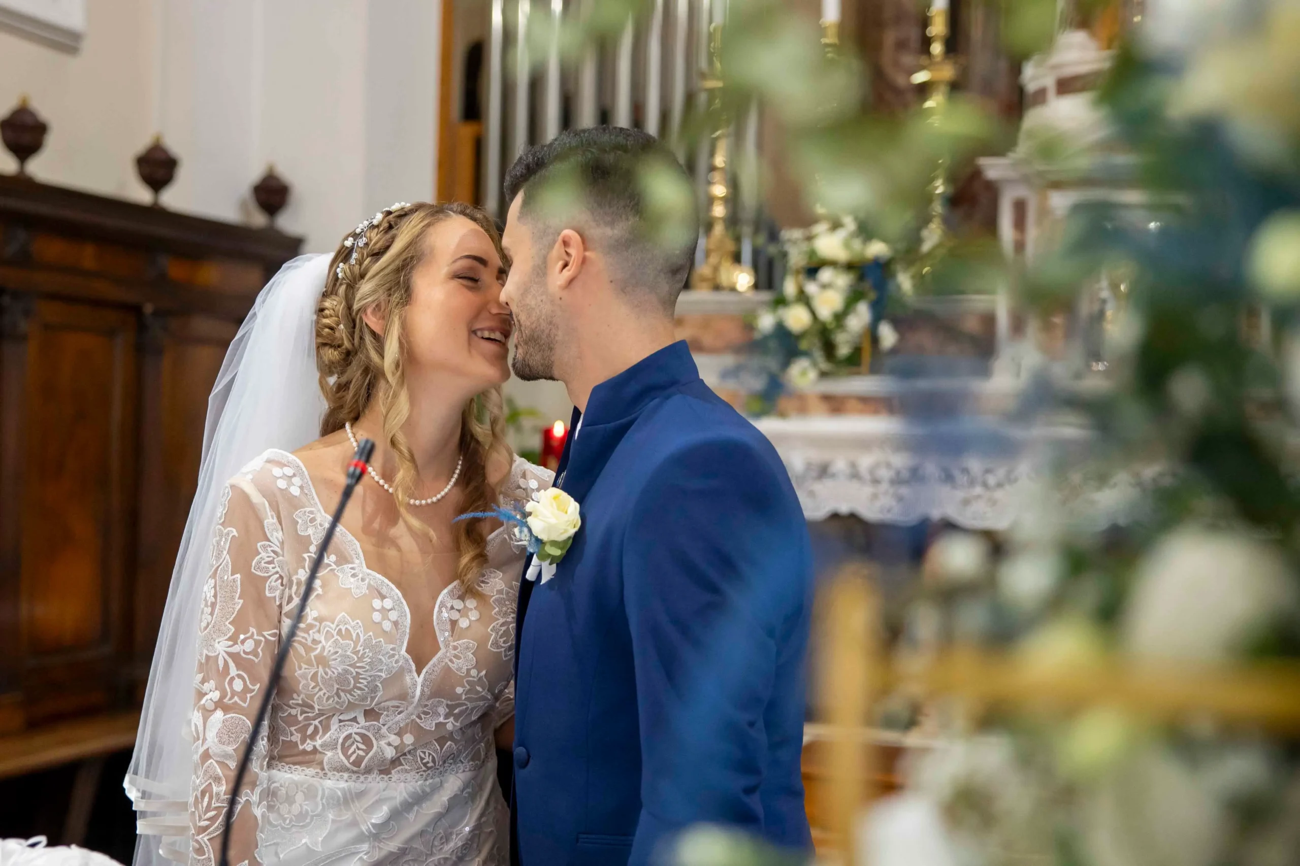 Primo piano di una sposa e uno sposo che si baciano all’interno di una chiesa durante la cerimonia nuziale, circondati da un’elegante decorazione di fiori e tessuti bianchi e blu che creano un’atmosfera romantica e solenne.