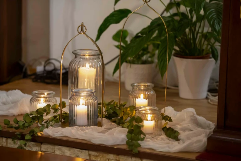 Primo piano di alcune candele accese all’interno di barattoli di vetro, poggiate su una superficie in legno chiaro, circondate da piante verdi rigogliose e tessuto bianco che ne completano l’atmosfera naturale e accogliente.