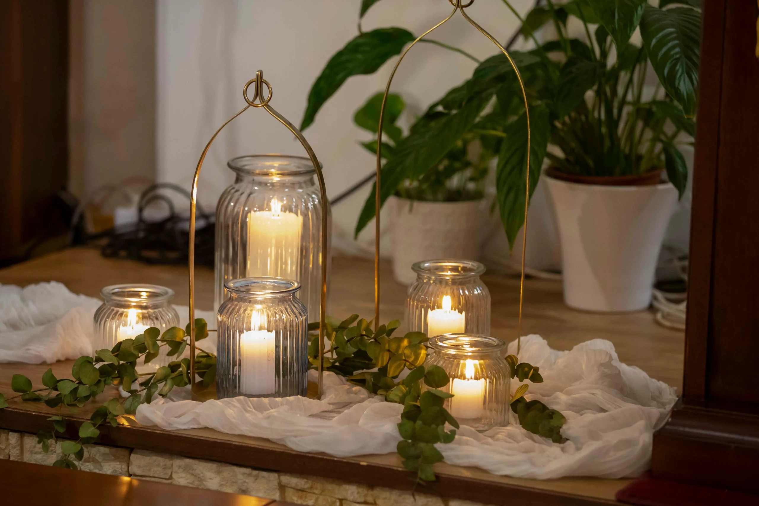 Primo piano di alcune candele accese all’interno di barattoli di vetro, poggiate su una superficie in legno chiaro, circondate da piante verdi rigogliose e tessuto bianco che ne completano l’atmosfera naturale e accogliente.