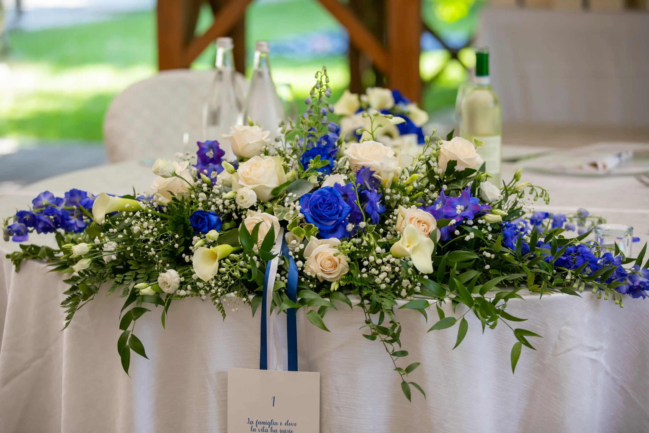 Elegante centrotavola per eventi, realizzato con un ricco mix di rose fresche bianche e blu, mescolate a fogliame verde decorativo e piccoli fiori bianchi di riempimento. La composizione floreale è appoggiata al centro di un tavolo per banchetti, coperto da una tovaglia bianca impeccabile.