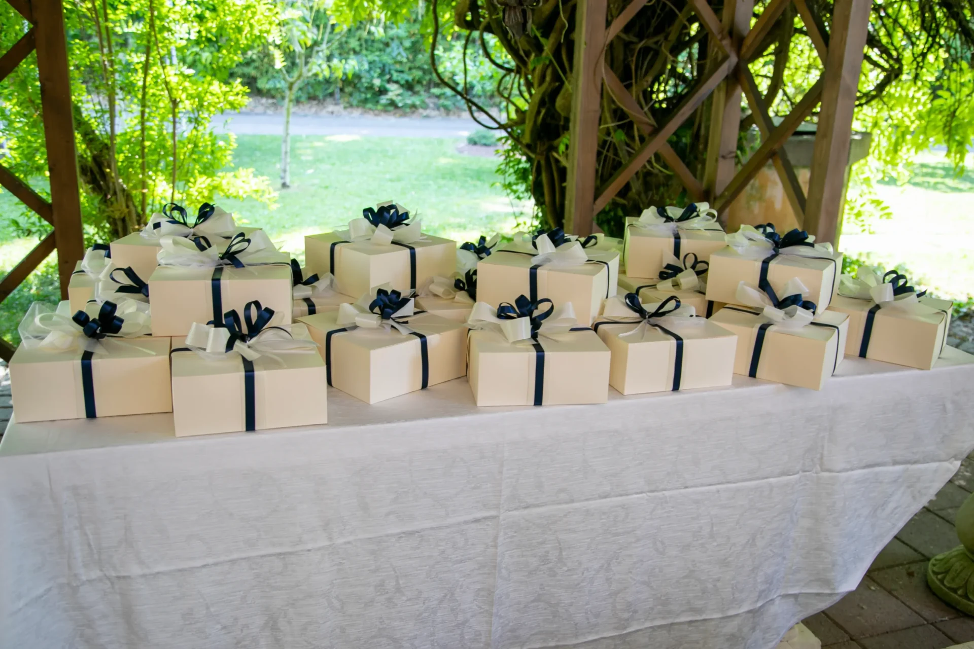 Tavolo apparecchiato in modo elegante, ricoperto da una tovaglia bianca finemente ricamata, con sopra numerose bomboniere di colore bianco legate con nastri di raso blu. La scena è allestita all'aperto, sotto una tettoia o gazebo di legno rustico