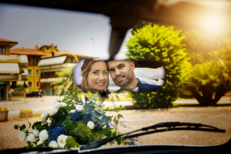 Riflesso unico e creativo di una coppia di sposi felici all'interno di un'auto, ripresi attraverso lo specchietto retrovisore. La vista mostra i loro volti sorridenti e, attraverso il parabrezza, il cruscotto e un mazzo di fiori adagiato sul cofano con l'ambiente esterno sullo sfondo.