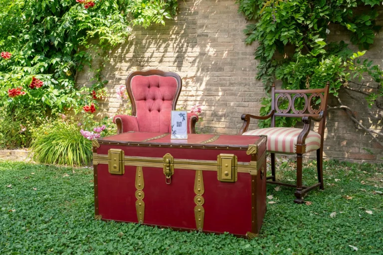 Baule rosso antico e due sedie da esterno moderne in vimini nel lussureggiante giardino di una villa storica, con un muro di pietra rossa rustica sullo sfondo. Perfetto per l'arredo da giardino di lusso e l'architettura paesaggistica.