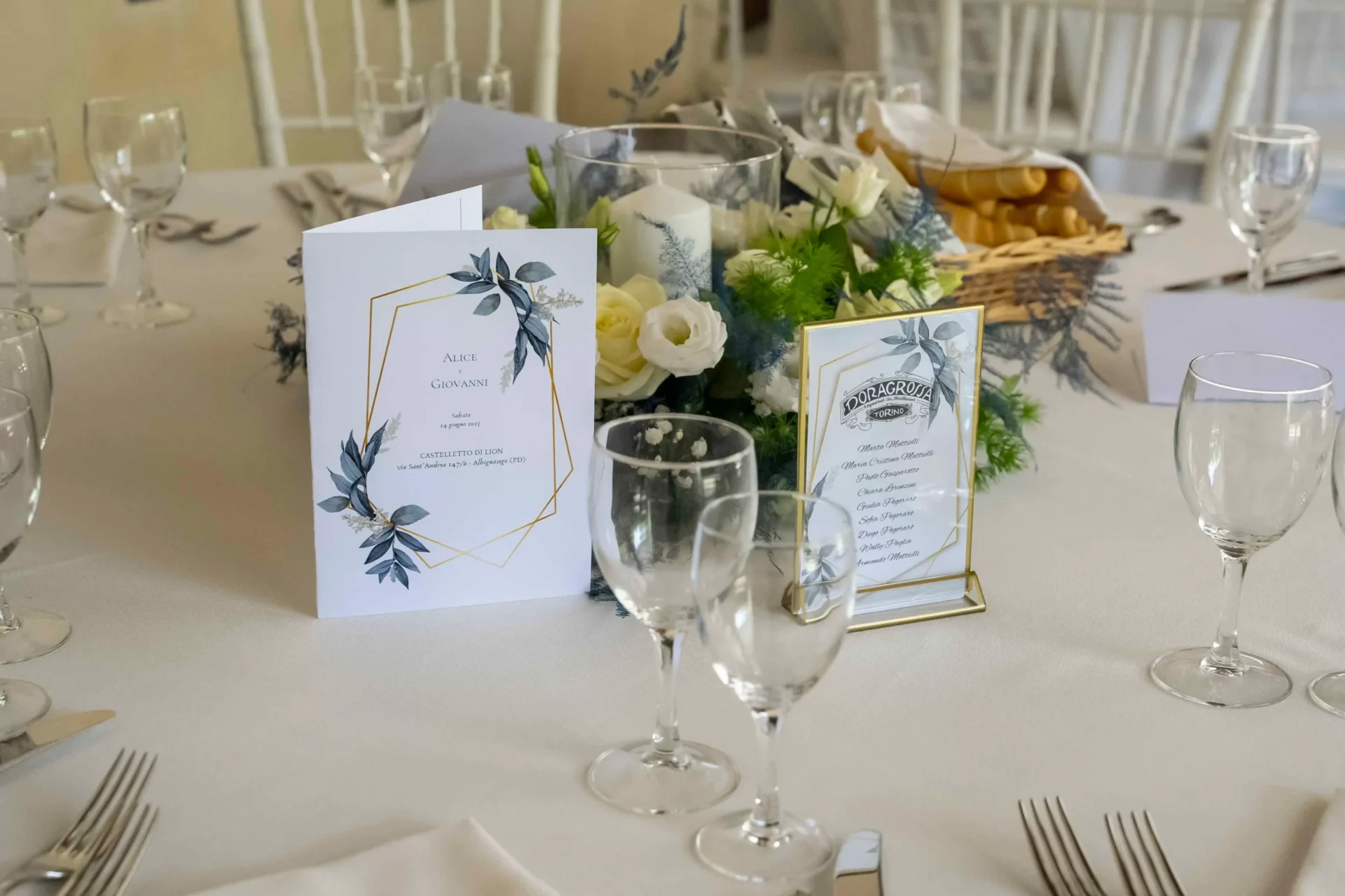 Primo piano di un tavolo da ricevimento elegantemente apparecchiato: spiccano un raffinato centrotavola con composizioni di fiori freschi, bicchieri in cristallo e posate. Ogni posto è contrassegnato da un piedistallo che sorregge un cartoncino con il nome degli invitati (segnaposto).