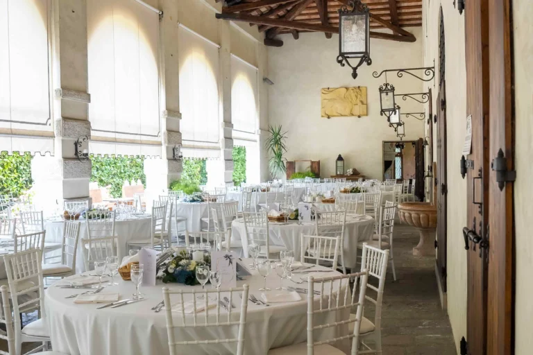 Ampia vista di un allestimento per matrimoni, che mostra una serie di tavoli elegantemente apparecchiati e sedie da cerimonia disposte attorno. La scena è ambientata all'aperto, protetta da un grande portico colonnato di una villa storica.