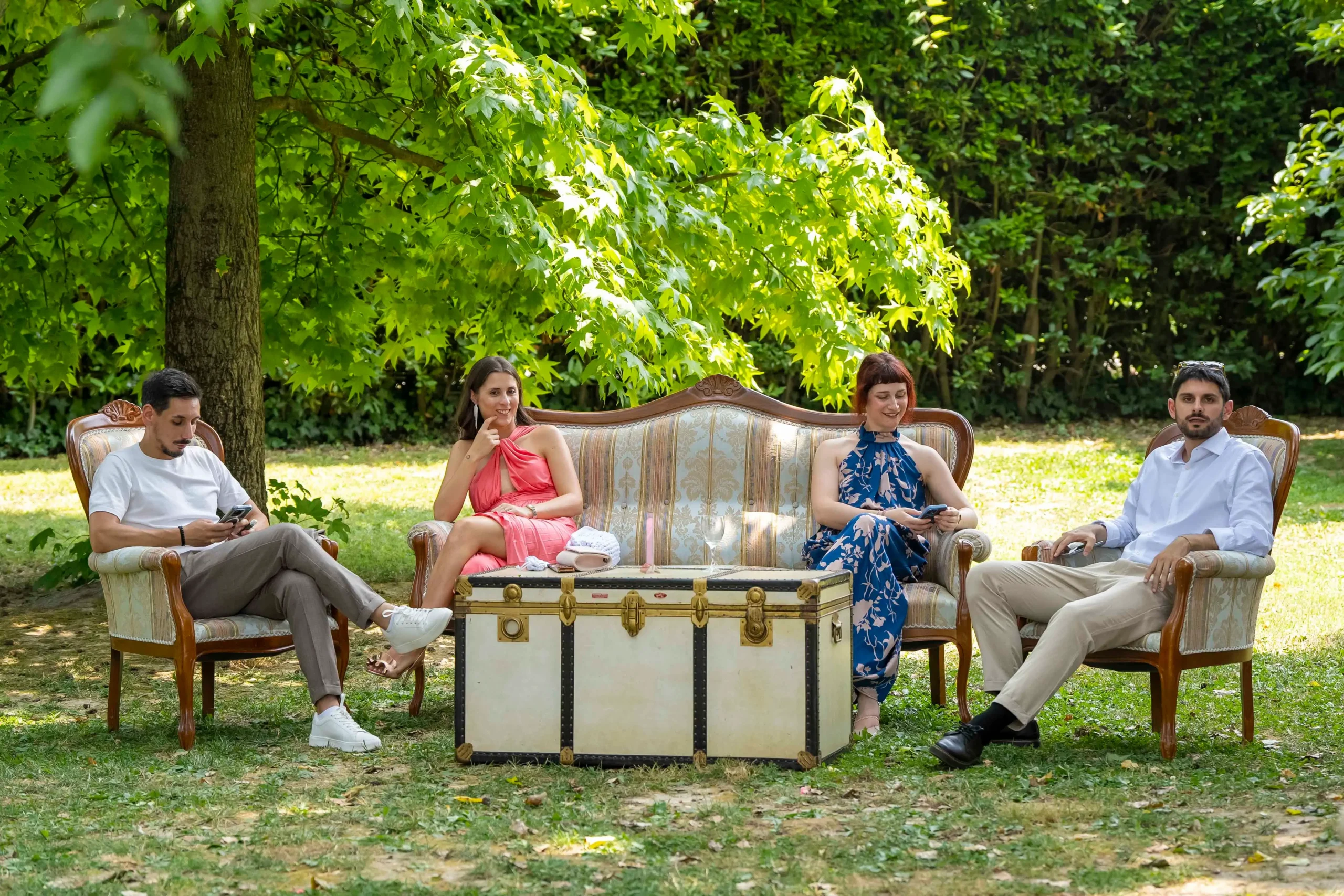 Persone in abiti eleganti sedute su divanetti e poltroncine vintage immersi nella natura e nel verde di un giardino