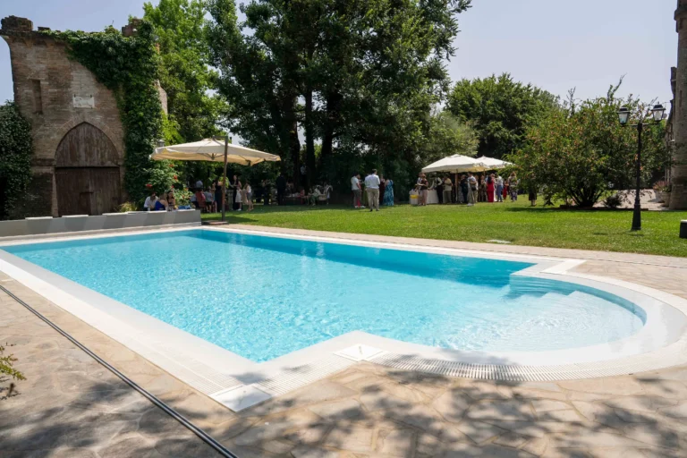 Ampia veduta di una lussuosa piscina esterna di una villa elegante. Nel giardino adiacente, invitati vestiti con abiti da cerimonia passeggiano e si intrattengono sotto ombrelloni che coprono i buffet allestiti. L'atmosfera generale è luminosa e suggestiva, documentando un momento vibrante di un ricevimento nuziale serale o pomeridiano all'aperto.