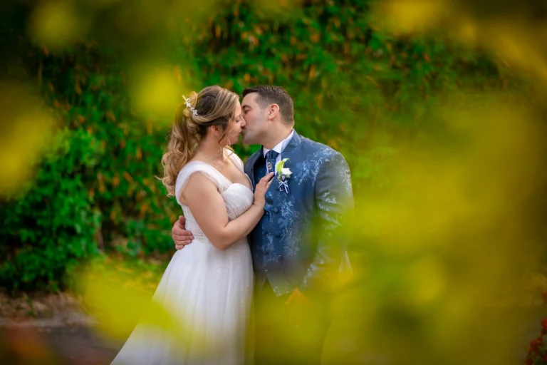 Una coppia di sposi, vestiti elegantemente con abito bianco e completo scuro, si scambia un tenero e romantico bacio. Sono incorniciati naturalmente da un arco o una fitta vegetazione di foglie verdi e rami delicati, creando un'atmosfera intima e fiabesca.