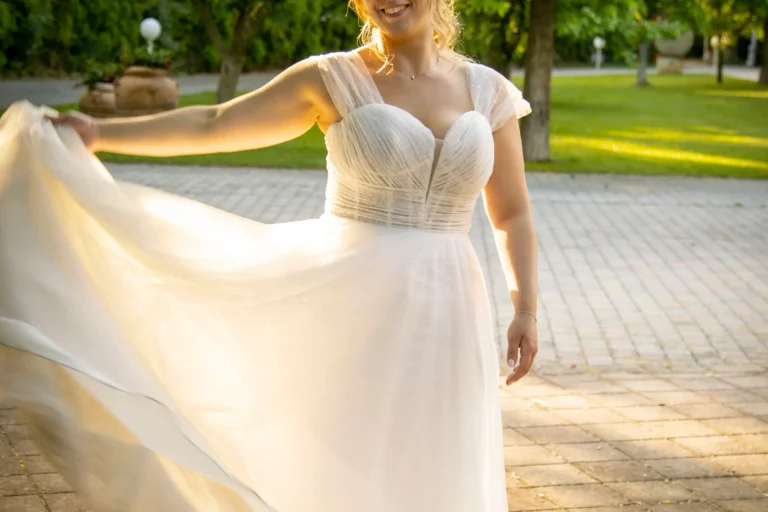 Primo piano di una sposa in un elegante e voluminoso abito bianco, in raso, in posa nel suo giorno di nozze. Sullo sfondo sfocato si estende un bellissimo giardino lussureggiante con alberi adulti e vegetazione curata.