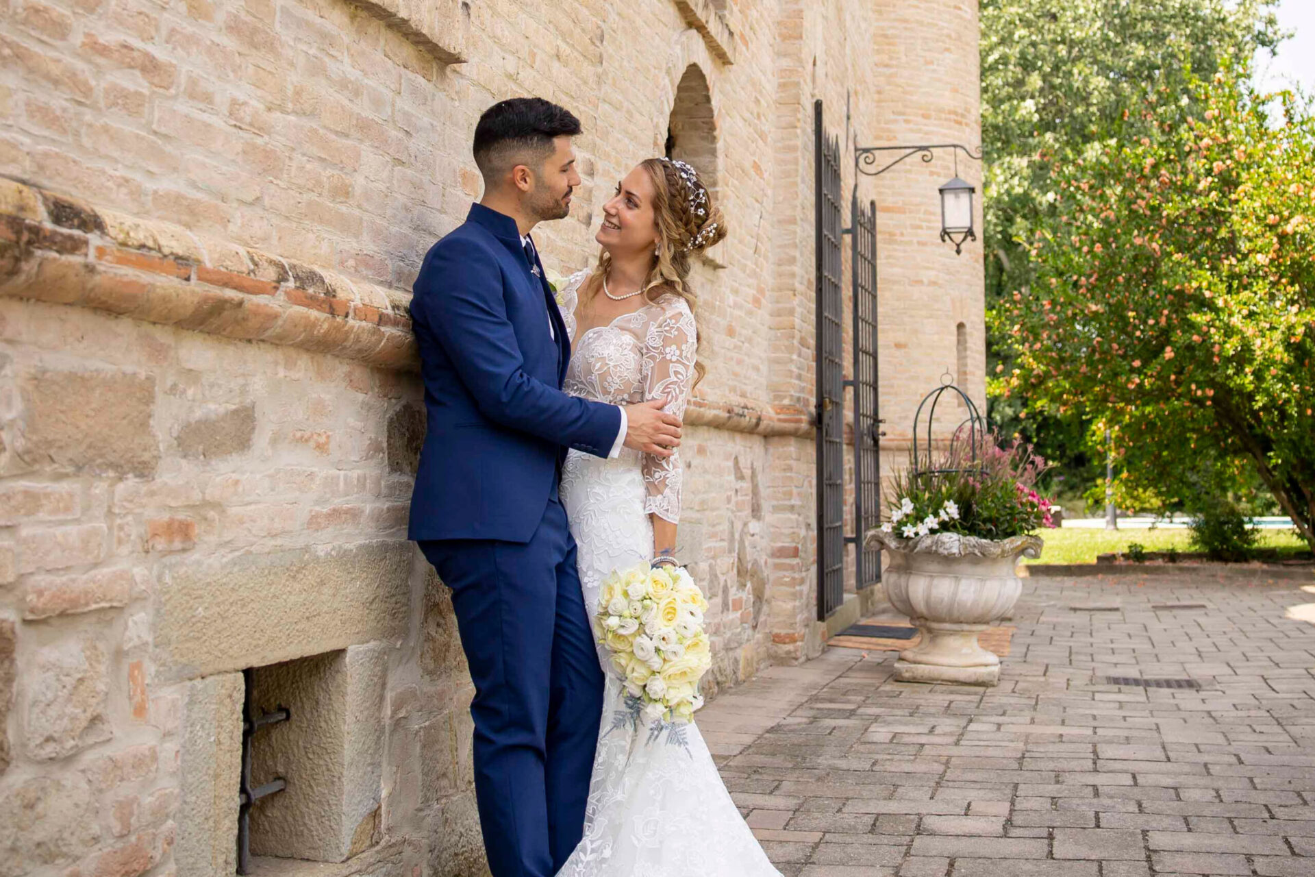 foto di due sposi ritratti mentre sono appoggiati a una parete di una dimora medioevale mentre si guardano a vicenda con sullo sfondo un giardino alberato.