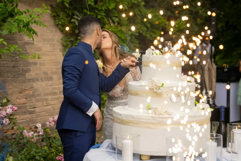 Una felice coppia di sposi, vestiti in abito da cerimonia, è ripresa mentre si scambia un tenero bacio durante il tradizionale taglio di una grande torta nuziale a più piani, riccamente decorata con crema bianca e delicate rose color pastello. Intorno a loro, zampilli luminosi di stelle filanti creano un'atmosfera magica, su uno sfondo di un suggestivo muro di mattoni rossi coperto da edera rampicante.