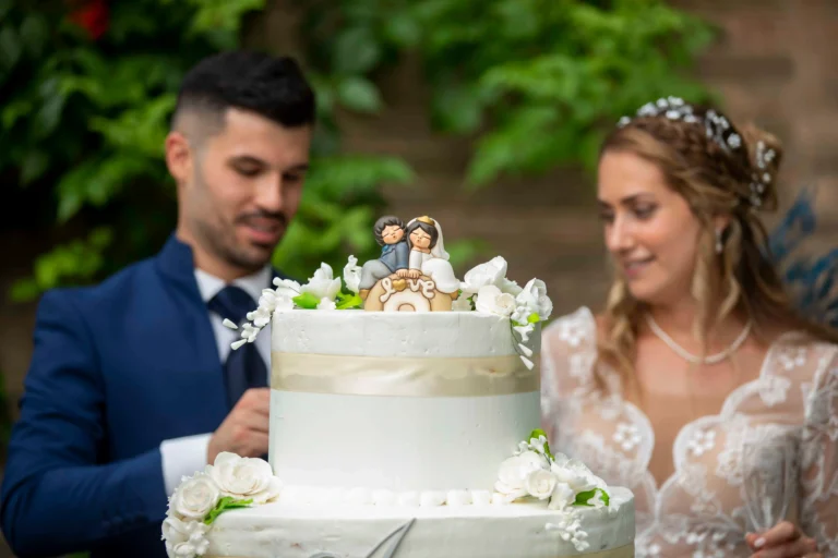 Primo piano di una torta nuziale a più piani, riccamente decorata con panna e composizioni di fiori bianchi freschi. Sulla cima è posizionata una statuina topper raffigurante due sposi in ceramica. Sullo sfondo sfocato, la sposa e lo sposo si preparano al tradizionale momento del taglio della torta
