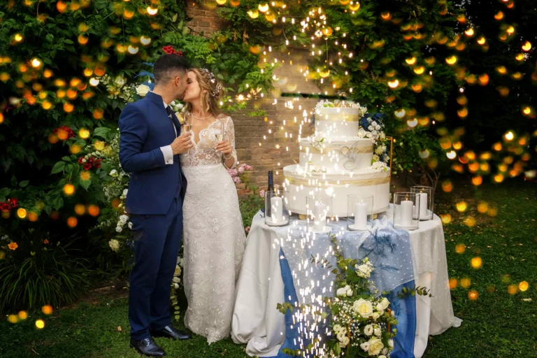Una felice coppia di sposi, vestiti in abito da cerimonia, è ripresa mentre si scambia un tenero bacio daccanto ad una grande torta nuziale a più piani, riccamente decorata con crema bianca e delicate rose color pastello. La torta poggia su un piccolo tavolino decorato con tovaglaito bianco e blu e un mazzo di rose e fiori freschi davanti. Intorno a loro, zampilli luminosi di stelle filanti creano un'atmosfera magica, su uno sfondo di un suggestivo muro di mattoni rossi coperto da edera rampicante.