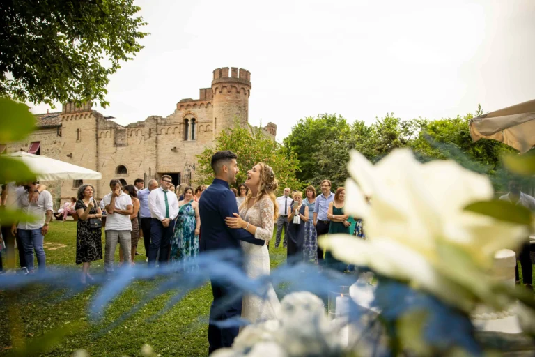 Ampia vista di un elegante giardino per ricevimenti, pieno di numerosi invitati vestiti a festa che osservano. Al centro della scena, la coppia di sposi danza felicemente, celebrando il loro primo ballo.