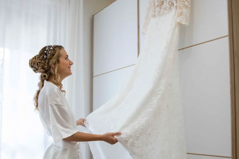 Una donna in primo piano, osserva con emozione il suo voluminoso abito da sposa bianco appeso all'armadio, durante il giorno del matromonio. Lei tiene delicatamente la parte bassa della gonna.