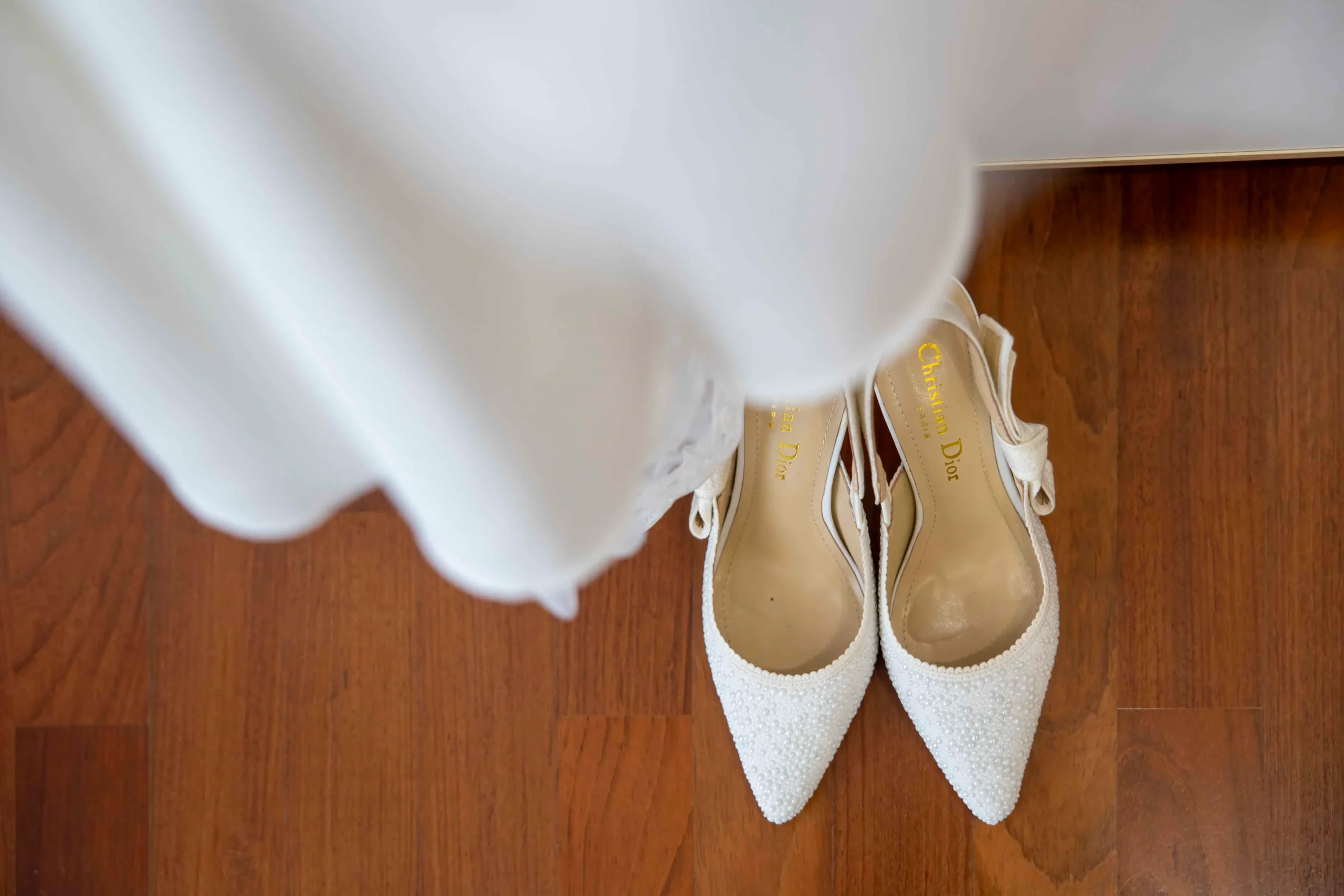 Primo piano di un paio di décolleté bianche di lusso decorate con perle, appoggiate su un pavimento di legno e parzialmente coperte dalla gonna dell’abito da sposa che ricade con eleganza sopra di esse.