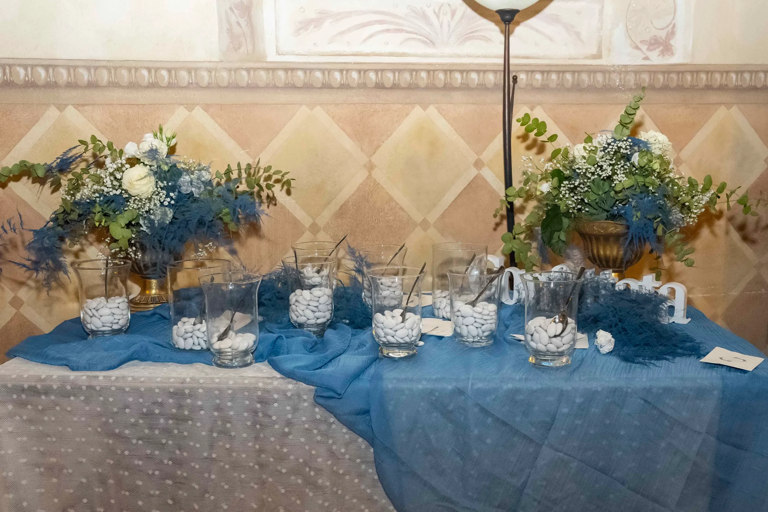 Vista di un tavolo tematico per la confettata , splendidamente addobbato e decorato per un matrimonio. Il tavolo è coperto da un tovagliato bianco e blu finemente ricamato e adornato con vasi di ottone contenenti mazzi di fiori freschi. Davanti una varietà di vasi di vetro espongono confetti di diversi gusti.