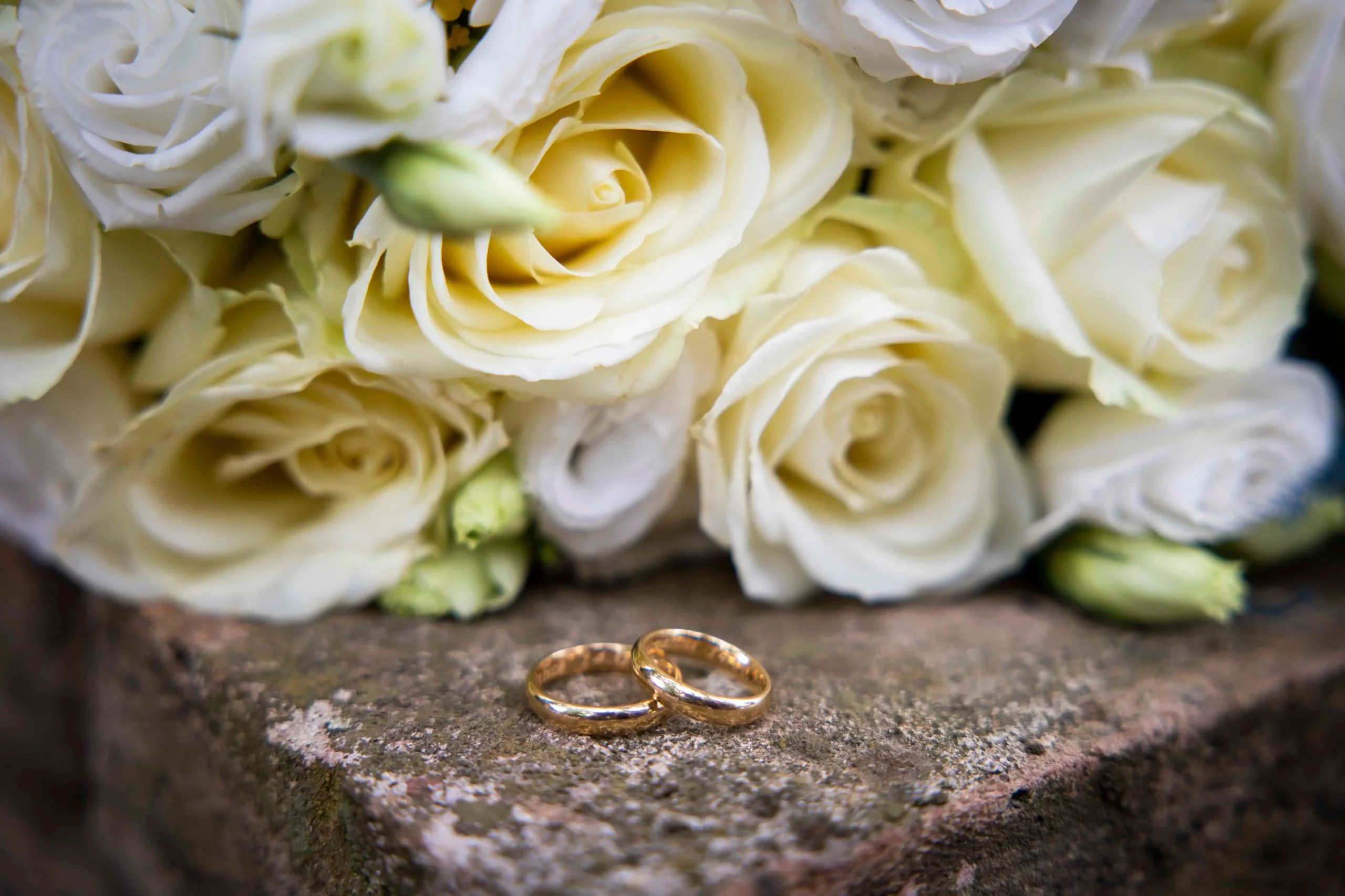 Primo piano di due fedi nuziali in oro appoggiate su una superficie di pietra e sullo sfondo un mazzo di rose bianche.