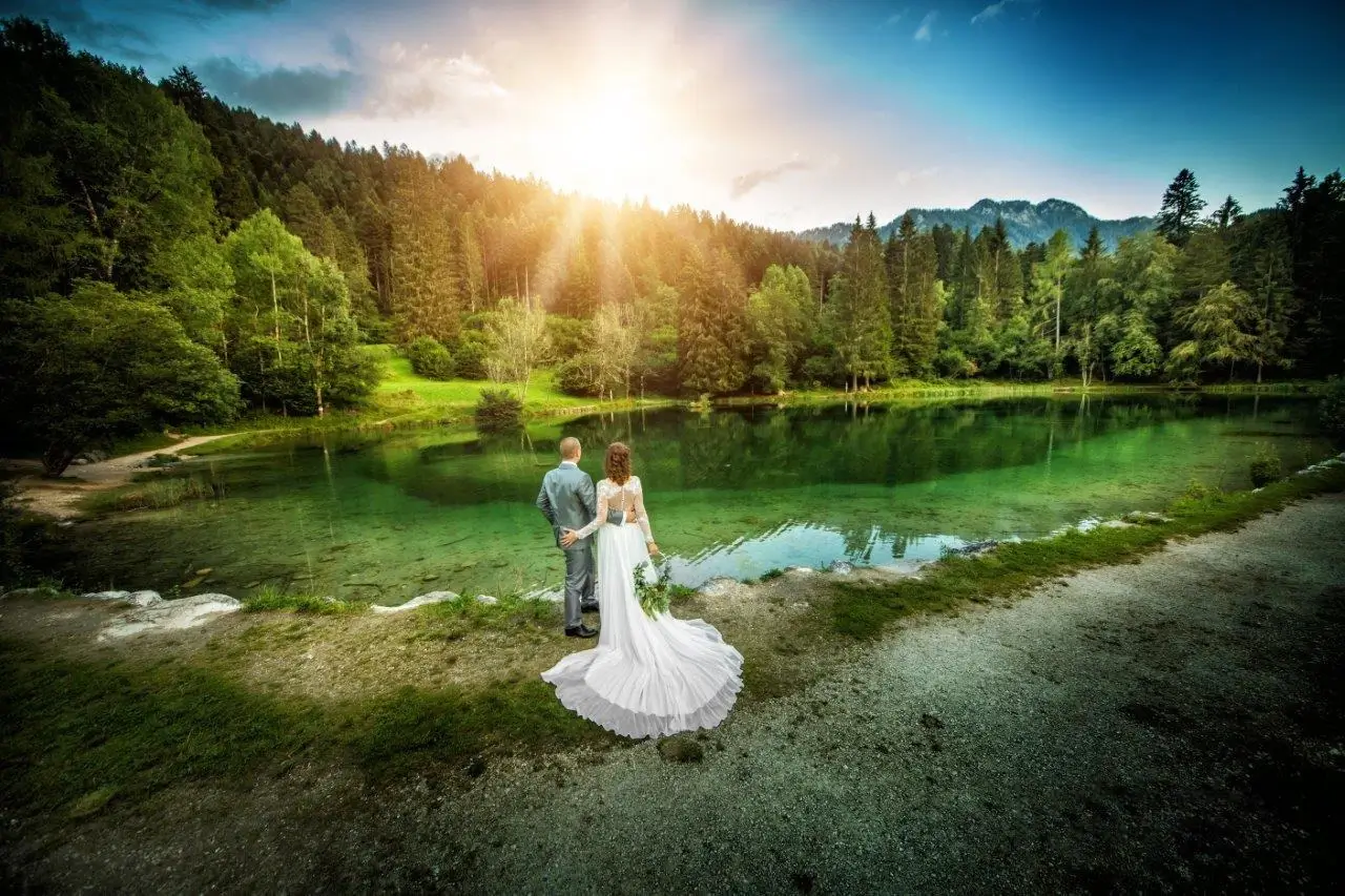Coppia di sposi di spalle, vestiti da matrimonio, che ammirano un idilliaco paesaggio alpino. In primo piano, un laghetto naturale con acqua limpida color smeraldo, circondato da fitti alberi verdi e maestose montagne sullo sfondo.