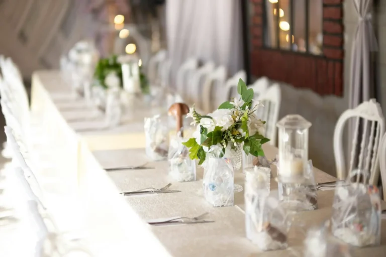 Vista di un tavolo da ricevimento nuziale, splendidamente apparecchiato e decorato interamente con elementi bianchi. Sono visibili una tovaglia immacolata, composizioni di fiori bianchi freschi e altre decorazioni chiare (come candele o vasi).