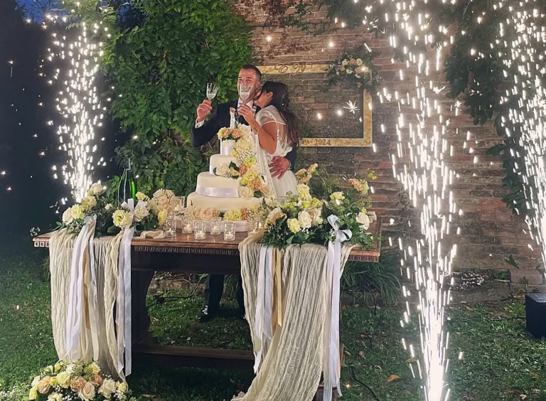 Una felice coppia di sposi, vestiti in abito da cerimonia, è ripresa mentre si scambia un tenero bacio durante il tradizionale taglio di una grande torta nuziale a più piani, riccamente decorata con crema bianca e delicate rose color pastello. Intorno a loro, zampilli luminosi di stelle filanti creano un'atmosfera magica, su uno sfondo di un suggestivo muro di mattoni rossi coperto da edera rampicante.